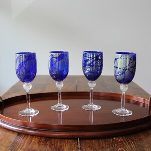 May include: Four blue wine glasses with abstract white and blue patterns are displayed on a dark wooden serving tray with brass handles. The glasses have clear stems and a decorative base.