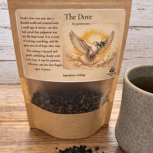 Könnte beinhalten: Ein brauner Papiersack mit „The Dove“ Oolong-Tee mit einem transparenten Fenster, einer Taubenillustration und Text. Eine graue Keramiktasse und lose Teeblätter liegen neben dem Beutel auf einer Holzoberfläche.