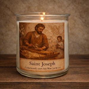 May include: A lit, clear glass candle jar with a label featuring an illustration of Saint Joseph. The label also includes the text "Saint Joseph", "Cedarwood | 100% Soy Wax | 30 hr 5 oz". The candle sits on a wooden surface.