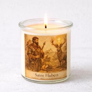 May include: A lit, clear glass candle jar with a label featuring an illustration of Saint Hubert, a man with a dog, a deer, and a cross. The candle is scented with sandalwood and saffron, and is 5 oz.