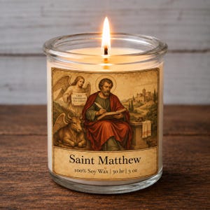 May include: A clear glass candle with a lit wick, featuring a label depicting Saint Matthew writing at a desk with an angel and an ox. The label includes the text Saint Matthew, 100% Soy Wax, 30 hr, and 5 oz, set against a vintage-style illustration.