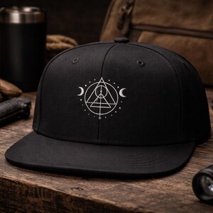 May include: Black baseball cap with a white geometric design featuring a triangle, circle, and crescent moons. The cap is on a wooden surface with a knife, gloves, and a flashlight in the background.