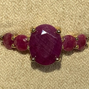 May include: A gold ring featuring a large, oval-cut ruby gemstone as the centerpiece, flanked by smaller, round rubies. The rubies are a deep, rich red color, and the ring has a classic, elegant design.