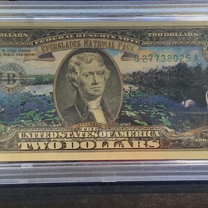 May include: A two-dollar bill featuring a portrait of Thomas Jefferson, with a scene of the Everglades National Park. The bill is encased in a protective holder, with text indicating it is authenticated and uncirculated.