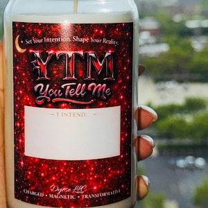 May include: A clear glass candle jar with a white wax fill and a central wick. The label features a red and black glittery background with the text "Set Your Intention. Shape Your Reality.", "YTM", "You Tell Me", "I INTEND...", and "Dyese LLC".
