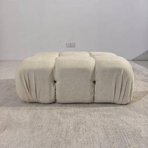 May include: A cream-colored, tufted ottoman with a rectangular shape. The ottoman has a soft, textured fabric and is divided into six cushioned sections. Dark buttons secure the sections. The piece is a neutral color and would fit in many home decor styles.