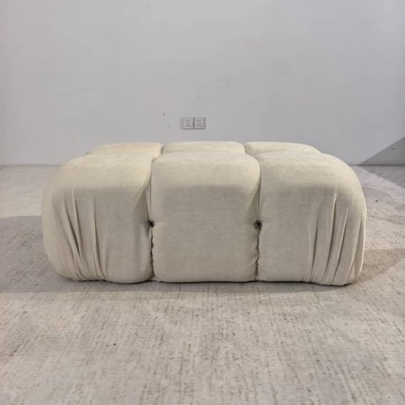 May include: A cream-colored, tufted ottoman with a rectangular shape. The ottoman has a soft, textured fabric and is divided into six cushioned sections. Dark buttons secure the sections. The piece is a neutral color and would fit in many home decor styles.