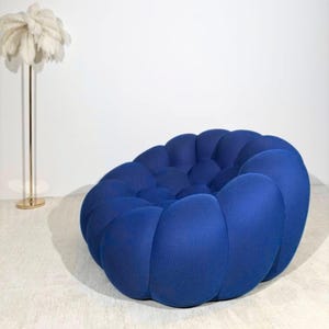 May include: A contemporary, circular blue armchair with a distinctive, textured pattern. The chair is a vivid shade of blue and has a series of rounded, connected sections. A gold floor lamp with a white feather-like shade is also visible.