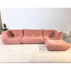 Op de afbeelding: Een roze corduroy modulaire bank met bijpassende poef. De bank heeft een laag profiel en een afgerond ontwerp. De stof heeft een geribbelde textuur. De poef is vierkant. De meubels staan op een lichtgekleurd tapijt.