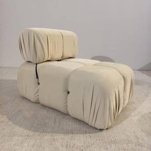 May include: A cream-colored, modular sofa with a rounded, upholstered design. The sofa features a low back and armrests, with a series of connected, cushioned sections. The fabric has a soft, velvety texture. The overall aesthetic is modern and comfortable.