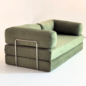 Puede incluir: Un sofá de pana verde con un diseño minimalista. El sofá presenta un perfil bajo, con una base rectangular y reposabrazos redondeados. Un marco de metal plateado añade un toque moderno. El sofá está sobre un fondo blanco.