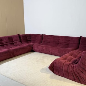 May include: A burgundy corduroy sectional sofa, arranged in an L-shape. The sofa has multiple sections with a quilted design on the cushions. It is positioned on a light-coloured carpet. The overall style is modern and comfortable.