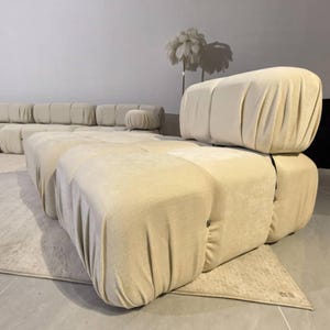 May include: Beige modular sofa with a rounded design. The sofa is composed of several large, upholstered blocks, creating a unique and modern aesthetic. The fabric has a soft, draped appearance. A small, round ottoman is also visible.