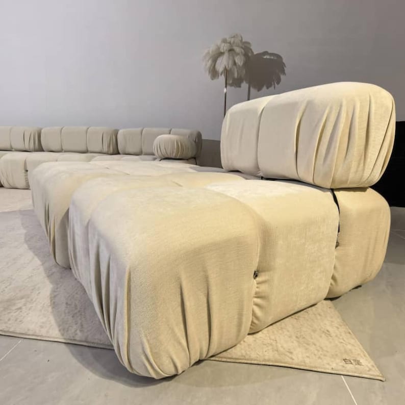 May include: Beige modular sofa with a rounded design. The sofa is composed of several large, upholstered blocks, creating a unique and modern aesthetic. The fabric has a soft, draped appearance. A small, round ottoman is also visible.