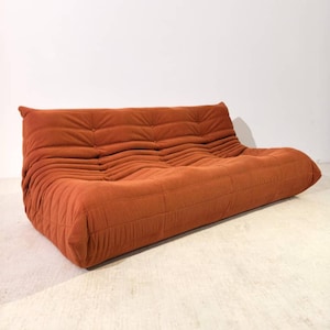 Op de afbeelding: Een oranje corduroy bank met een laag profiel en een gecapitonneerd ontwerp. De bank heeft een afgeronde vorm en een gestructureerd oppervlak, met een reeks horizontale ribbels over de zitting en rugleuning. De bank is een mid-century modern stijl.