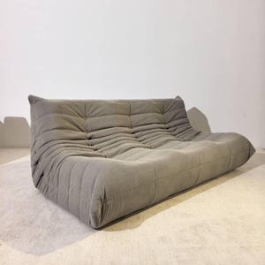 May include: A low-profile, multi-seat sofa upholstered in a textured, taupe-colored fabric. The piece features a distinctive, segmented design with horizontal tufted channels across the backrest and seat, creating a soft, rounded silhouette.