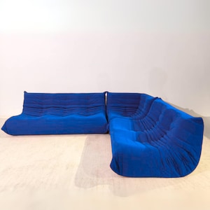 Może przedstawiać: Nowoczesna, modułowa sofa narożna w żywym niebieskim odcieniu. Sofa ma unikalny, teksturowany design z serią poziomych grzbietów na poduszkach. Zestaw dwuczęściowy jest ustawiony w kształcie litery L, oferując dużą przestrzeń do siedzenia.