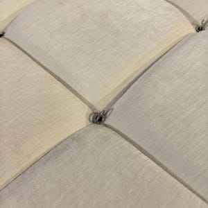 May include: Close-up of a section of a cream-colored, diamond-tufted fabric. The fabric is divided into diamond shapes by stitched lines. Small, silver-colored metal rings are at the intersections of the lines.