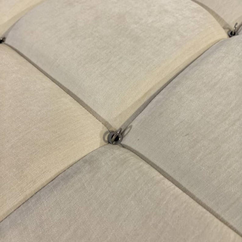 May include: Close-up of a section of a cream-colored, diamond-tufted fabric. The fabric is divided into diamond shapes by stitched lines. Small, silver-colored metal rings are at the intersections of the lines.