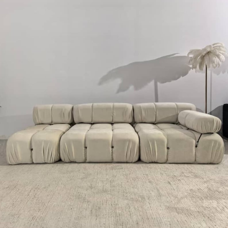 May include: A modular, cream-colored sofa with a low profile. The sofa is composed of several rectangular sections, with a rounded bolster pillow on the right. The fabric has a soft, velvety texture. The sofa sits on a neutral-toned rug.