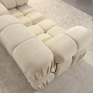 May include: A modular, cream-colored sofa with a unique, rounded design. The sofa is composed of multiple cushioned sections, creating a textured, inviting surface. The neutral color and modern design make it a versatile piece for any living space.