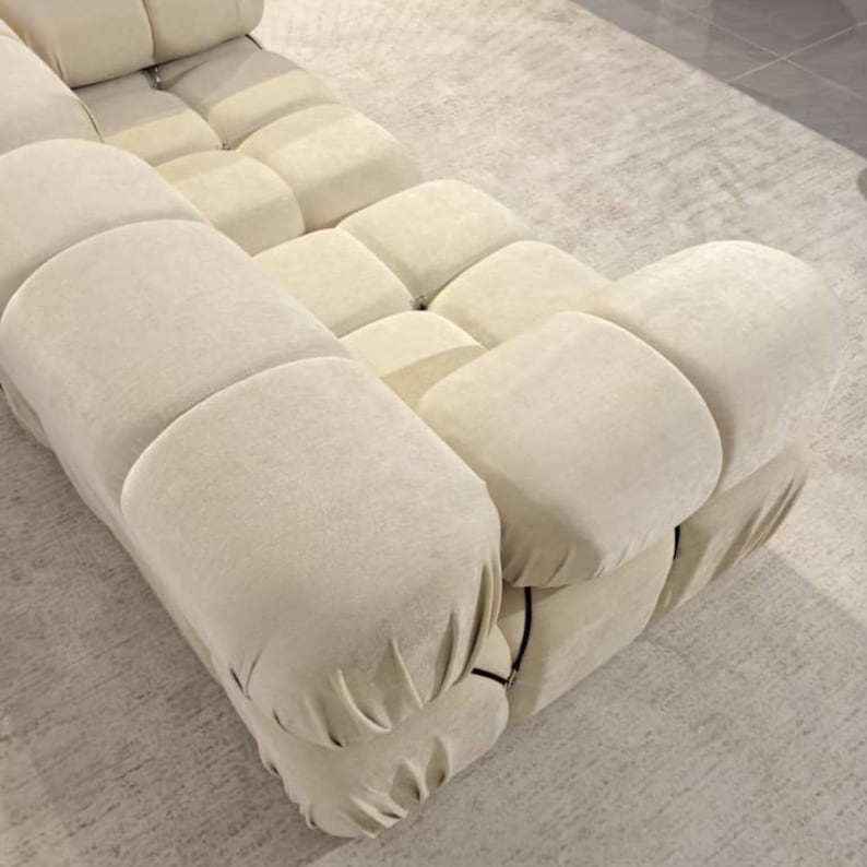 May include: A modular, cream-colored sofa with a unique, rounded design. The sofa is composed of multiple cushioned sections, creating a textured, inviting surface. The neutral color and modern design make it a versatile piece for any living space.