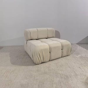 May include: A cream-colored, modular sofa with a rounded, blocky design. The sofa is upholstered in a soft, textured fabric and features a low profile. The design includes a separate backrest cushion, and the overall aesthetic is modern and minimalist.