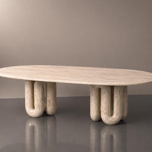 Op de afbeelding: Een lichtbeige ovale salontafel. De tafel heeft een glad blad en wordt ondersteund door drie sets gebogen, U-vormige poten. De tafel is gemaakt van een steenachtig materiaal.