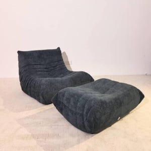 May include: A dark charcoal gray upholstered lounge chair and matching ottoman set. The chair features a low-profile, segmented design with horizontal tufted detailing across the backrest and seat, creating a soft, rounded, and plush appearance.