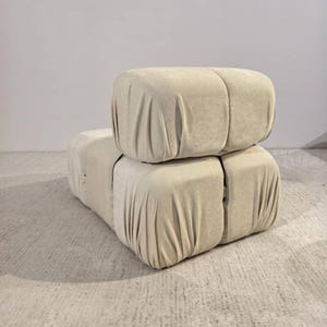 May include: A beige modular sofa with a soft, draped fabric. The sofa is composed of two stacked, cube-shaped sections, with visible seams and a neutral color palette. The design suggests a modern, minimalist aesthetic.