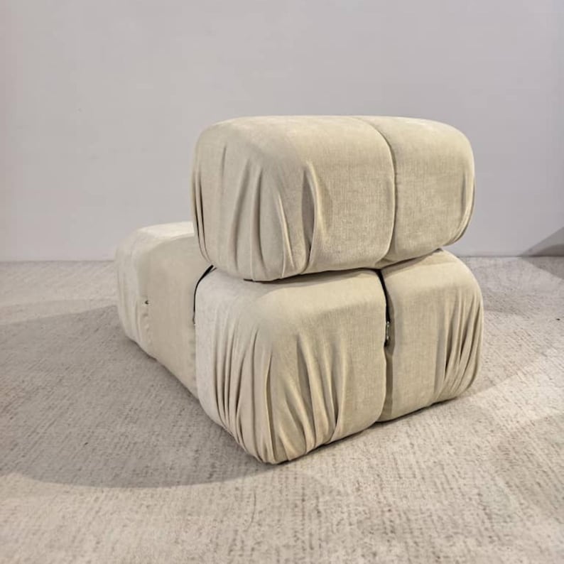 May include: A beige modular sofa with a soft, draped fabric. The sofa is composed of two stacked, cube-shaped sections, with visible seams and a neutral color palette. The design suggests a modern, minimalist aesthetic.