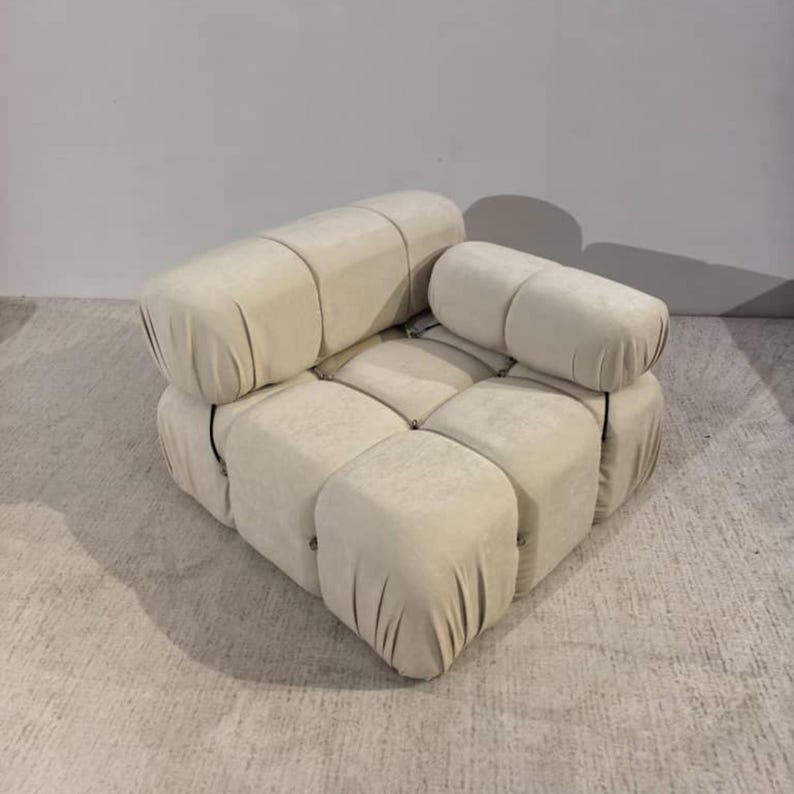 May include: A modular, cream-colored sofa with a unique design. The sofa is composed of several square and cylindrical cushions, arranged to form a comfortable seating area. The fabric has a soft, velvety texture. The design is modern and stylish.