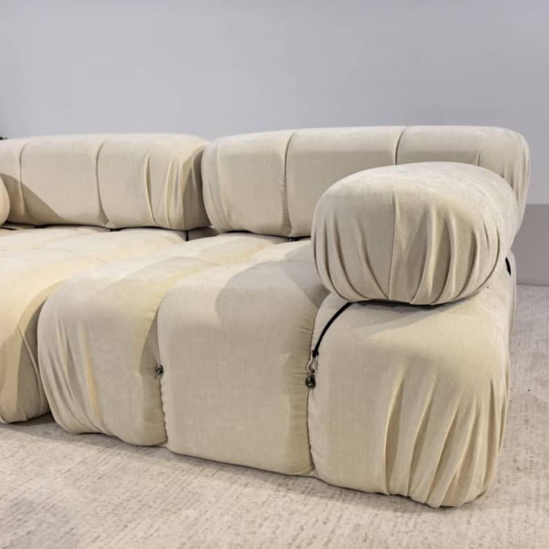 May include: A modular, cream-colored sofa with rounded, upholstered sections. The sofa features a soft, velvety texture and a modern design. The armrests are also rounded and covered in the same fabric. The sofa is placed on a light-colored rug.