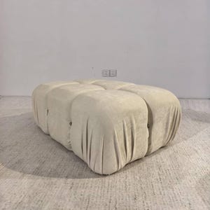May include: A large, beige ottoman with a tufted design. The ottoman has a rectangular shape with rounded edges and is covered in a soft, textured fabric. The design features multiple sections, creating a cushioned appearance.