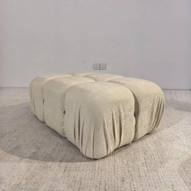 May include: A large, beige ottoman with a tufted design. The ottoman has a rectangular shape with rounded edges and is covered in a soft, textured fabric. The design features multiple sections, creating a cushioned appearance.