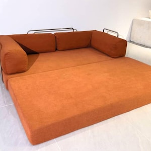May include: An orange sectional sofa with a pull-out bed feature. The sofa has a modern design with a rectangular shape and metal armrests. The cushions are a solid orange color, and the bed is fully extended.