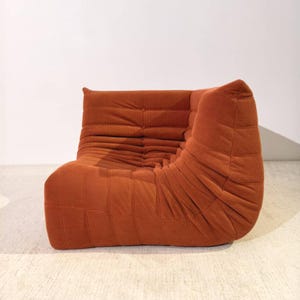 Può includere: Una poltrona in velluto a coste color arancione bruciato con un profilo basso e una forma arrotondata. La poltrona presenta schienale e seduta trapuntati, con un dettaglio a bottone. Il tessuto ha una trama a coste e la poltrona è su uno sfondo neutro.