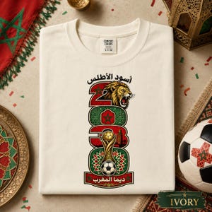 May include: An ivory t-shirt featuring a graphic design with the text 2023, a lion head, a gold trophy, and a soccer ball. The design incorporates the Moroccan flag colors of red and green with geometric patterns and Arabic script text.