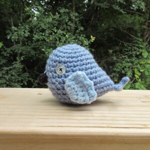 May include: A handmade, light blue crochet bird with a button eye and a small, darker blue beak. The bird has a light blue wing and tail, and is sitting on a wooden surface. The background is a blurred view of green foliage.