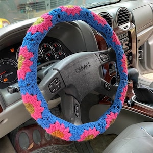 May include: A blue, pink, and yellow crocheted steering wheel cover. The cover is made of yarn and features a floral pattern. The cover is on a black steering wheel in a GMC vehicle. The interior of the car is visible.