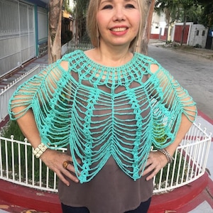 Puede incluir: Un poncho de crochet turquesa con un diseño de tejido abierto único. El poncho presenta un cuello redondo y flecos en las mangas y el dobladillo. Esta prenda hecha a mano es una adición elegante a cualquier guardarropa.