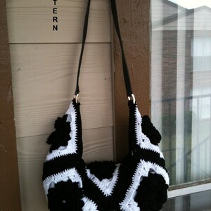May include: Black and white crocheted handbag with a black strap. The bag is made of squares with black flowers in the center. The bag is hanging on a hook.