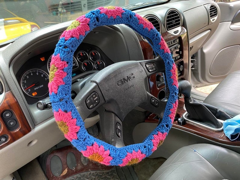 May include: A blue, pink, and yellow crocheted steering wheel cover. The cover is made of yarn and features a floral pattern. The cover is on a black steering wheel in a GMC vehicle. The interior of the car is visible.
