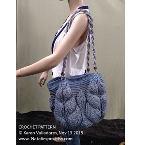 May include: A crocheted tote bag with a blue and gray color gradient. The bag has a rope handle and a textured, cable knit design. The bag is being held by a mannequin.