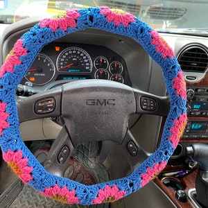 May include: A blue, pink, and yellow crocheted steering wheel cover. The cover is made of yarn and features a floral pattern. The cover is on the steering wheel of a GMC vehicle.