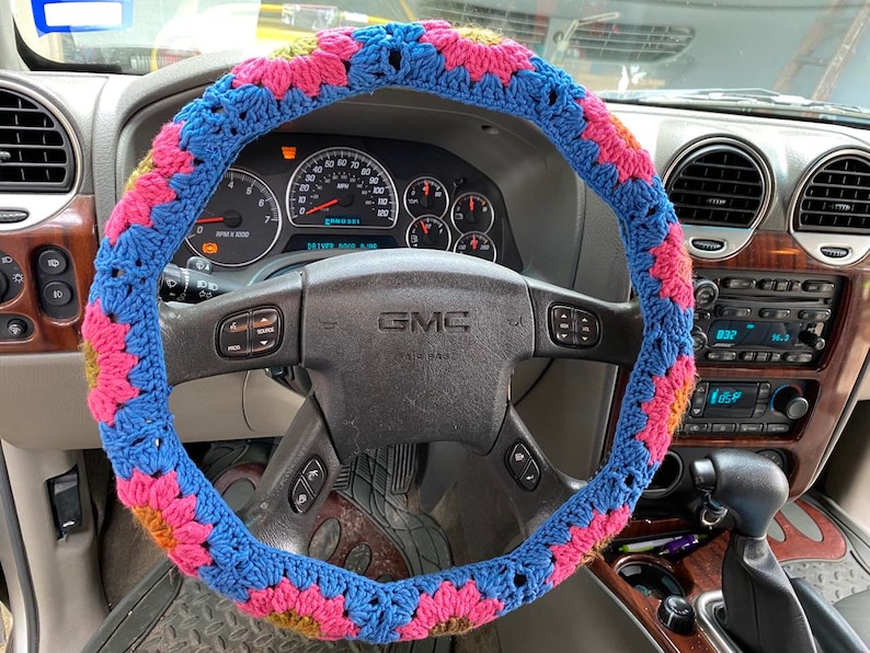 May include: A blue, pink, and yellow crocheted steering wheel cover. The cover is made of yarn and features a floral pattern. The cover is on the steering wheel of a GMC vehicle.