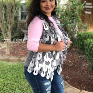 Puede incluir: Una mujer lleva un chaleco de ganchillo gris y blanco con una camisa rosa y pantalones vaqueros azules. El chaleco tiene un diseño festoneado y se lleva abierto. La mujer sonríe y mira a la cámara.