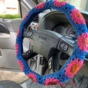 May include: A blue, pink, and yellow crocheted steering wheel cover. The cover is made of yarn and features a floral pattern. The steering wheel is black and has the GMC logo.