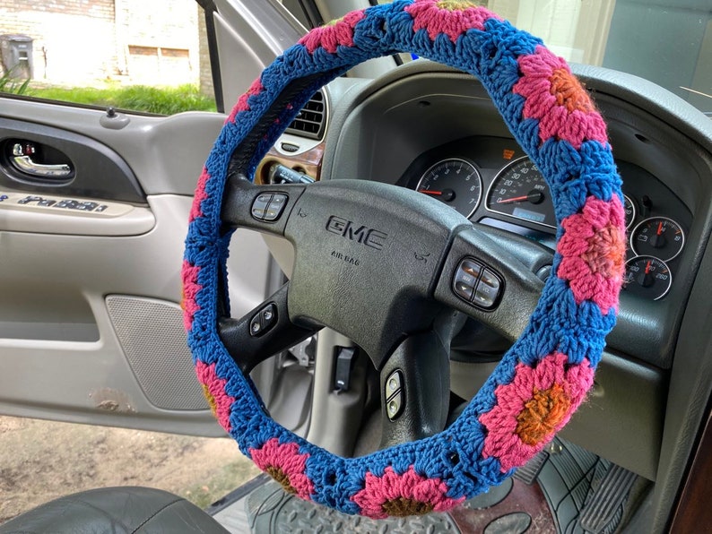 May include: A blue, pink, and yellow crocheted steering wheel cover. The cover is made of yarn and features a floral pattern. The steering wheel is black and has the GMC logo.