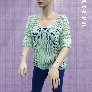 Peut inclure: Un haut en crochet vert clair avec un col en V et des manches courtes. Le haut pr&eacute;sente un motif ajour&eacute; d&eacute;licat et de petites fleurs en crochet autour du col et des manches. Le haut est port&eacute; par un mannequin sur un fond violet.